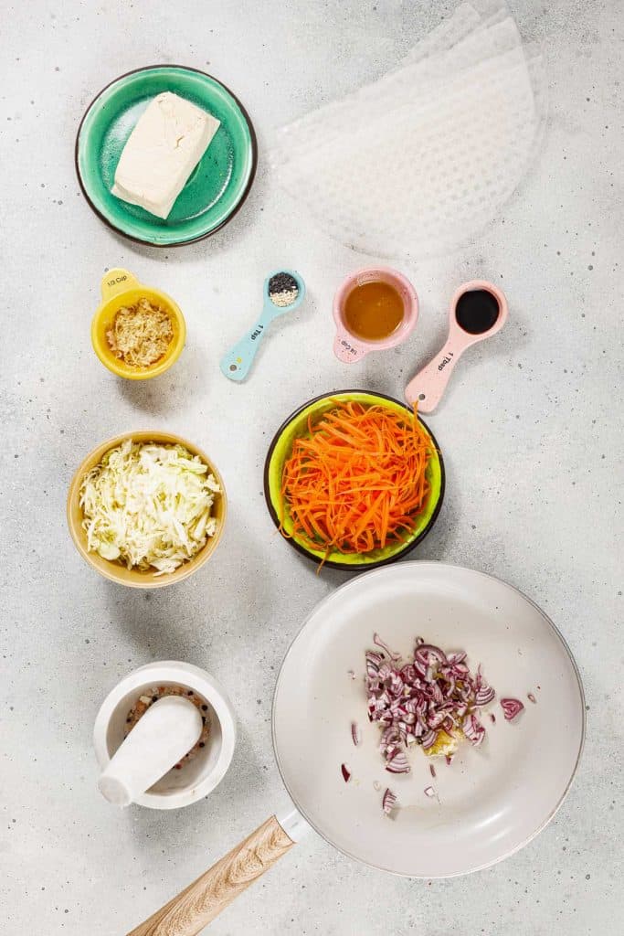 Flat lay of ingredients for spring rolls, including tofu, rice paper, shredded cabbage, carrot, ginger, seasonings, sauces, and a pan with chopped onions on a light gray surface.