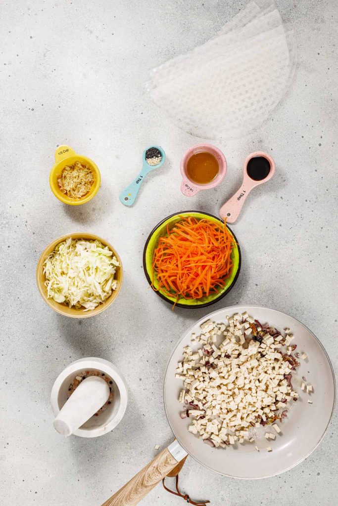 Overhead view of ingredients for spring rolls, including cabbage, carrots, tofu in a pan, ginger, seasoning, sauces in measuring cups, peppercorns in a mortar, and rice paper sheets.