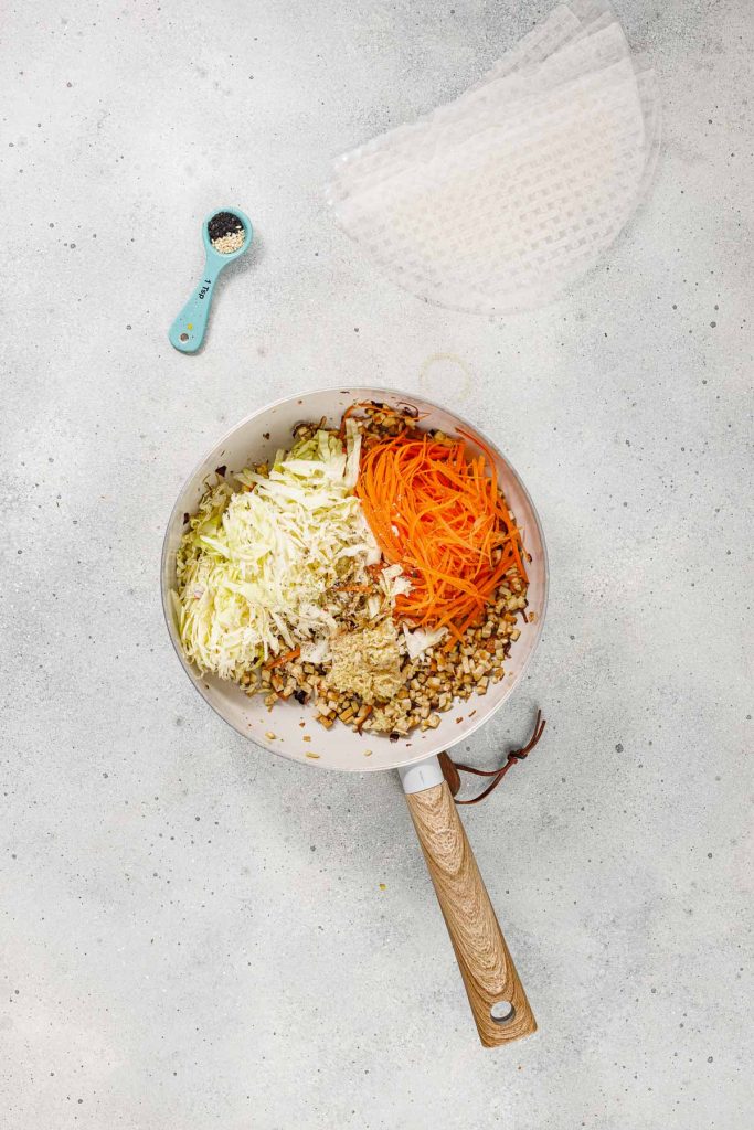 A pan with shredded cabbage, grated carrots, chopped mushrooms, and minced garlic; a spoon with pepper and rice paper sheets are nearby on a light countertop.