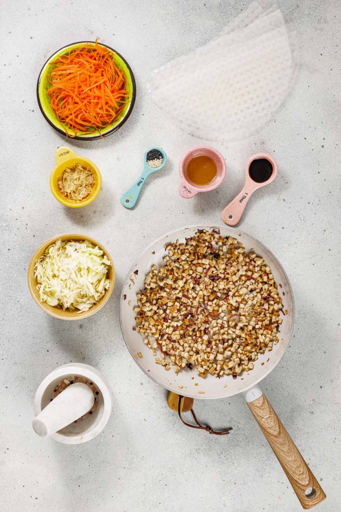 Top-down view of a pan with sautéed vegetables, bowls of shredded carrots, cabbage, ginger, and seasonings, with rice paper wrappers and a mortar and pestle on a white surface.