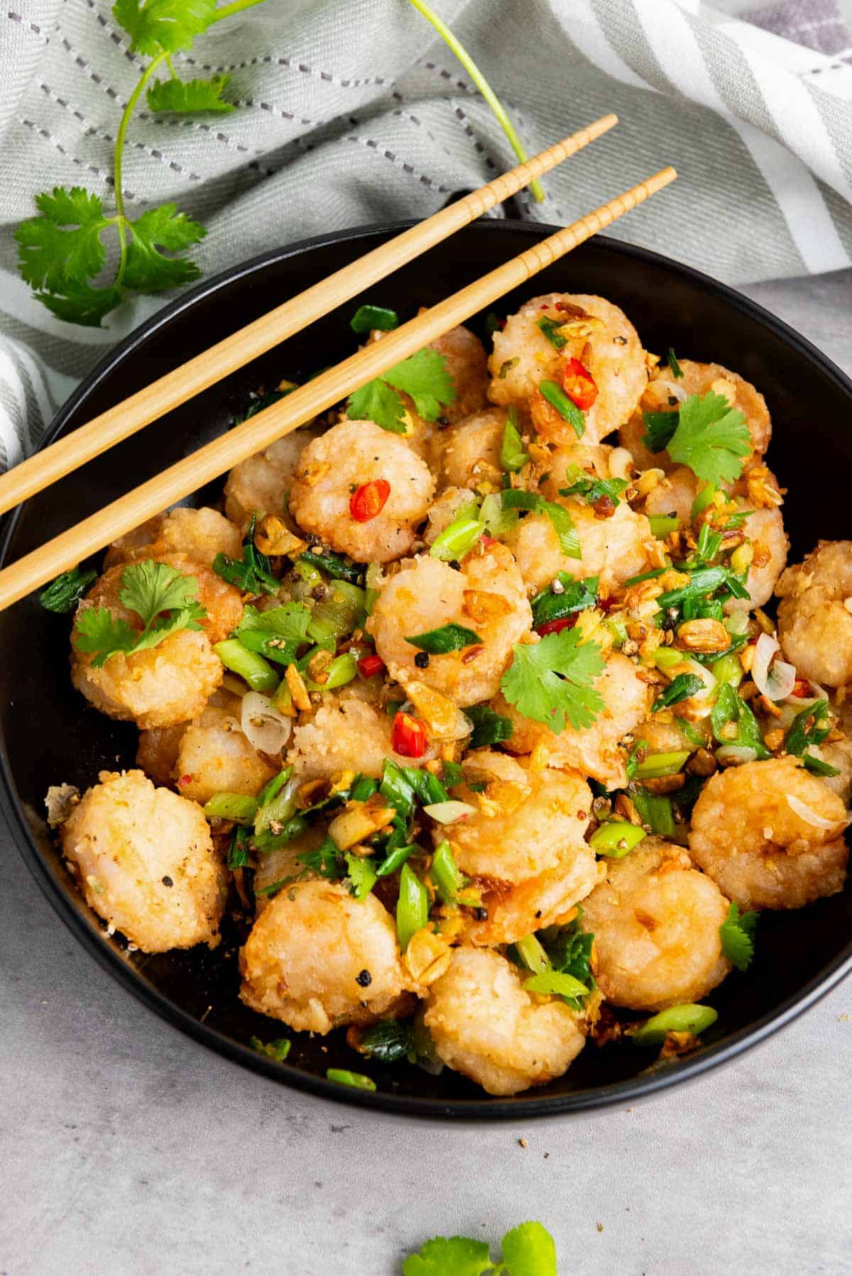 A black bowl filled with stir-fried shrimp garnished with chopped green onions, cilantro, red chili slices, and garlic, with wooden chopsticks resting on top.