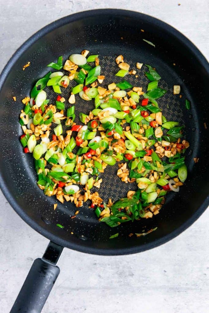 Chopped green onions, red chili, and garlic sautéing in a black frying pan on a gray surface.
