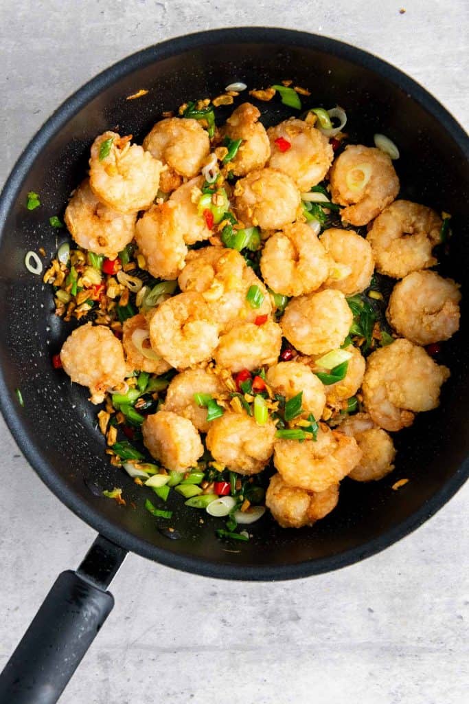 A black frying pan contains battered shrimp stir-fried with chopped green onions, red chili, and garlic on a light gray surface.