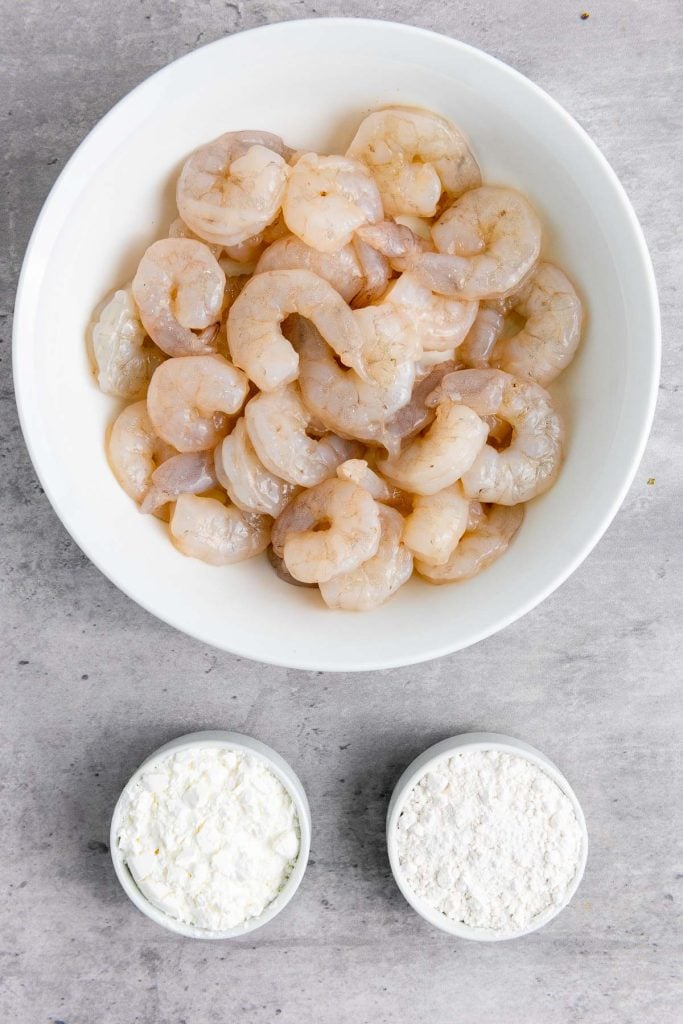 A white bowl of raw, peeled shrimp next to two small bowls filled with cornstarch and flour on a gray surface.