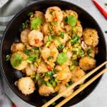 A black bowl filled with fried shrimp garnished with chopped green onions, red chili slices, garlic, and cilantro, with a pair of wooden chopsticks resting on the bowl.
