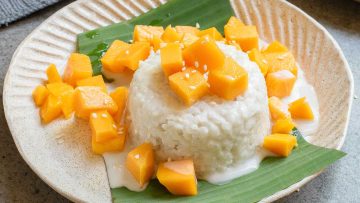 A plate of mango sticky rice with cubed mango, sweet coconut sauce, and sesame seeds, served on a green leaf.