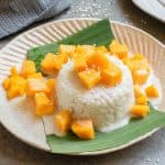 A serving of sticky rice topped with coconut cream, diced mango, and sesame seeds, presented on a banana leaf on a beige plate.