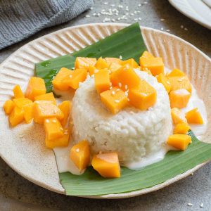 A serving of sticky rice topped with coconut cream, diced mango, and sesame seeds, presented on a banana leaf on a beige plate.