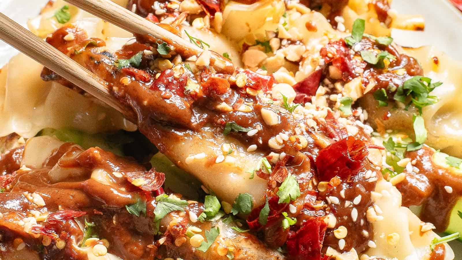 Close-up of dumplings drizzled with a rich sauce, topped with chopped peanuts, chili flakes, and fresh herbs. Chopsticks are picking up one dumpling, showcasing the vibrant toppings and textures.