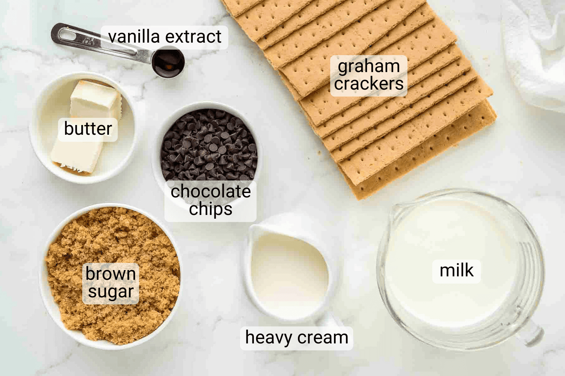 Ingredients for a recipe are arranged on a counter: graham crackers, chocolate chips, brown sugar, butter, milk, heavy cream, and vanilla extract.