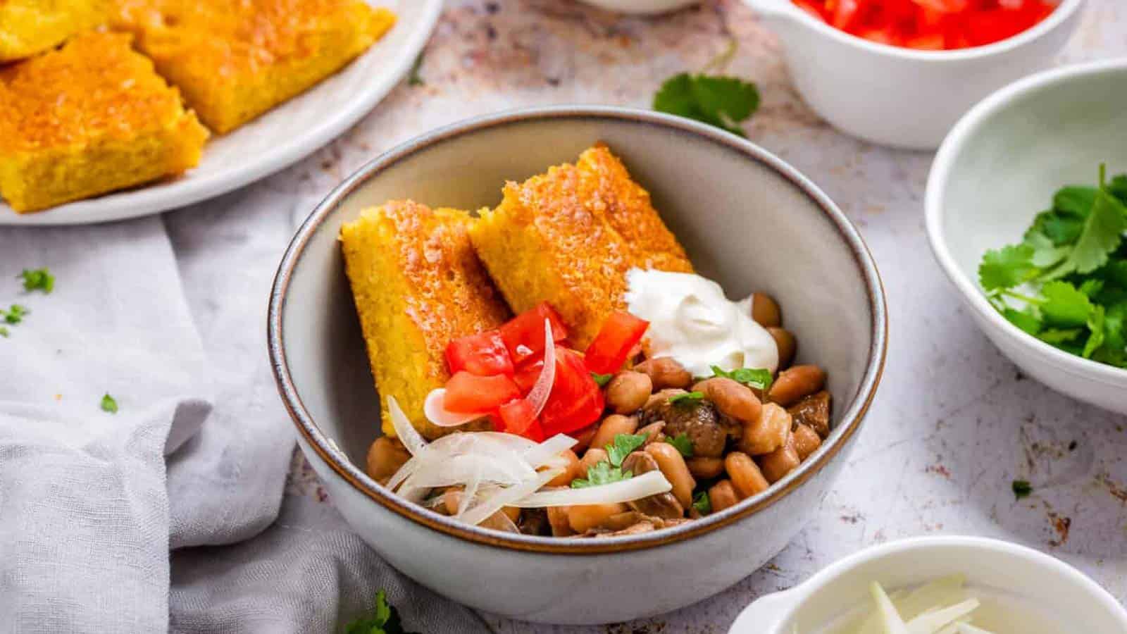 A bowl with cornbread, pinto beans, sour cream, diced tomatoes, and sliced onions. Surrounding bowls hold cornbread, chopped tomatoes, and fresh cilantro.
