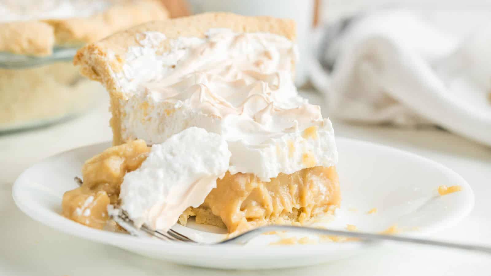 A slice of pie with a golden crust, creamy filling, and a layer of fluffy meringue on top sits on a white plate with a fork.