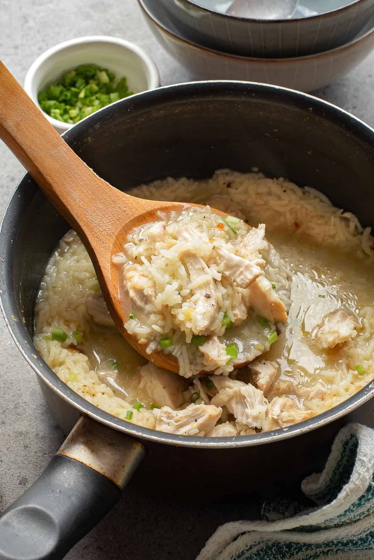 A saucepan with chicken and rice soup; a wooden spoon lifts a portion, and a small bowl of chopped green onions is nearby.