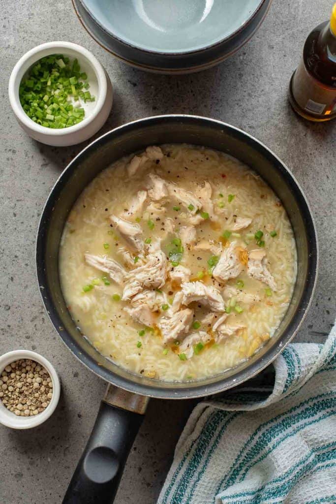 A pot of chicken and rice soup garnished with chopped green onions sits on a countertop next to bowls of pepper, green onions, and a striped towel.