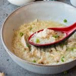 A bowl of chicken congee garnished with chopped green onions, with a red spoon lifting some rice and chicken from the broth.