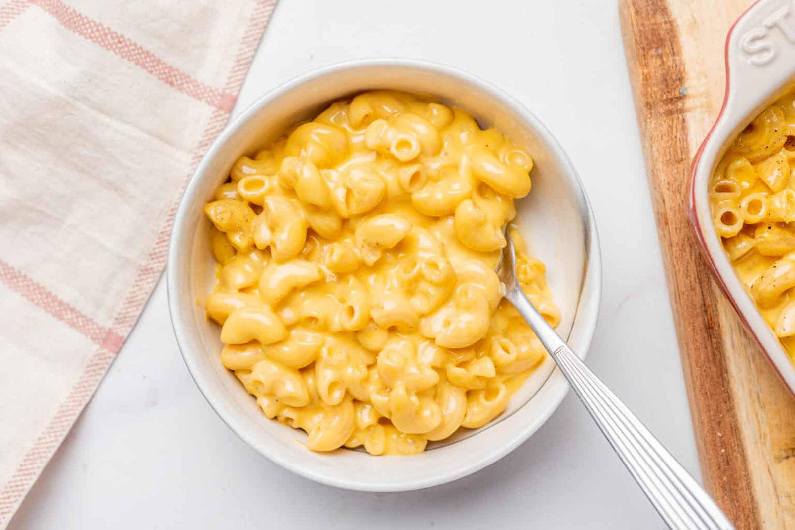 A bowl of macaroni and cheese with a fork, sitting on a white surface next to a towel and a wooden board.