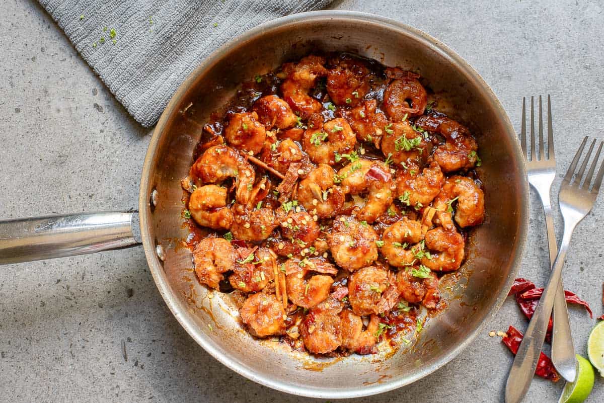 A stainless steel pan filled with cooked, saucy shrimp garnished with chopped herbs sits on a gray countertop next to two forks and a cloth napkin.