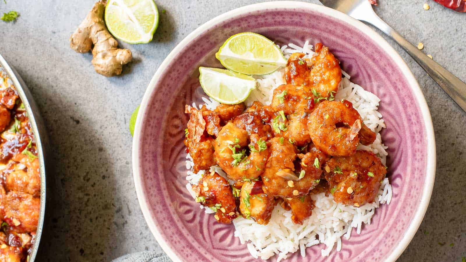 A bowl of white rice topped with saucy shrimp, garnished with lime wedges and herbs, sits on a gray surface next to ginger and a fork.