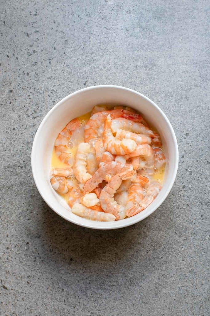 A white bowl filled with peeled, uncooked shrimp sitting in a light liquid on a gray textured surface.