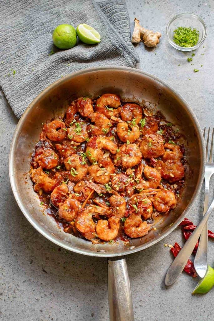 A stainless steel skillet filled with cooked shrimp in a brown sauce, garnished with chopped herbs, sits on a gray countertop with limes, ginger, and red chili peppers nearby.