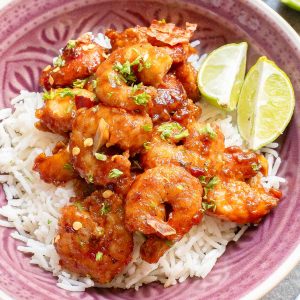 A bowl of white rice topped with spicy glazed shrimp, garnished with chopped herbs and served with two lime wedges.