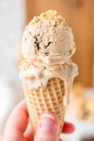 A hand holds an ice cream cone with two scoops of ice cream on top, one scoop slightly melting and dripping down the cone.