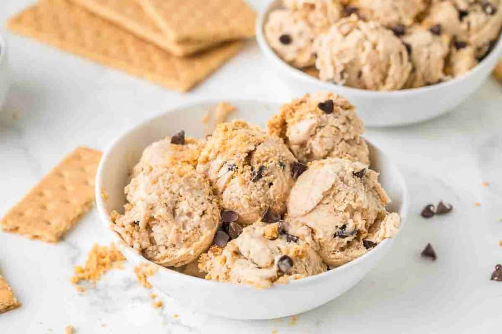 A white bowl filled with scoops of cookie dough ice cream, garnished with chocolate chips and graham cracker crumbs. Graham crackers and another bowl are visible in the background.