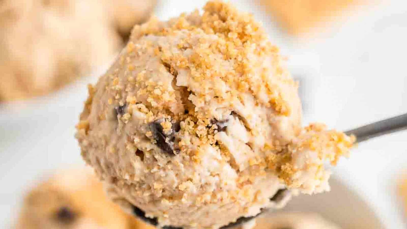 A close-up of a spoon holding a scoop of ice cream topped with a crumbly, brown topping, with blurred ice cream scoops in the background.