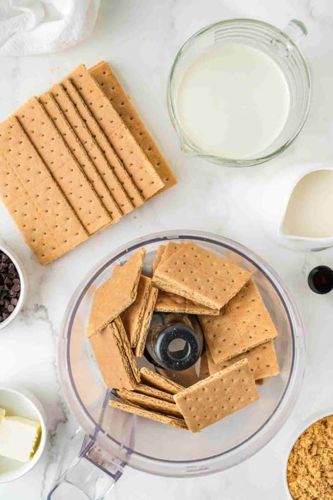 An overhead view of graham crackers in a food processor, with more crackers, a bowl of milk, butter, chocolate chips, and vanilla extract nearby on a white surface.