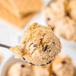 A close-up of a spoon holding a scoop of creamy dessert topped with crumbled graham crackers, with more dessert blurred in the background.