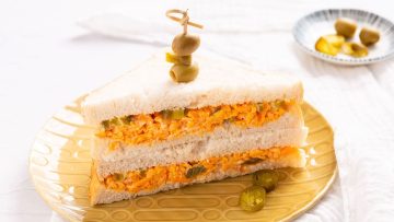 Two triangular halves of a sandwich filled with a shredded orange mixture, garnished with sliced olives on top, served on a patterned yellow plate with extra jalapeños on the side.