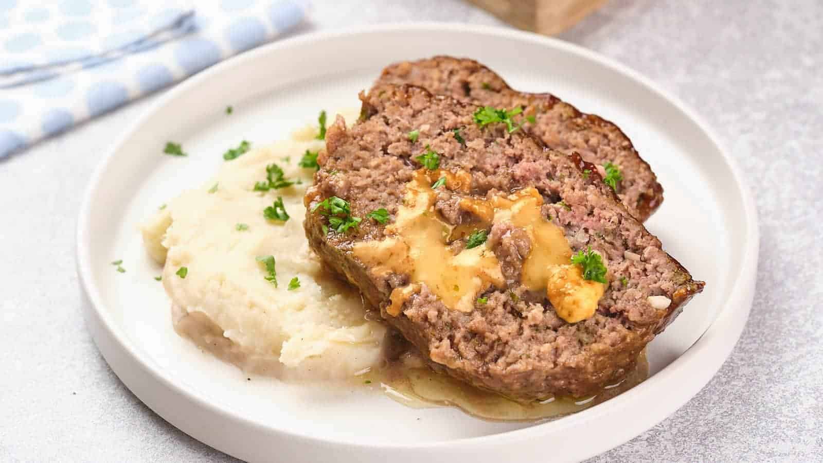 A plate with two slices of meatloaf topped with gravy, served alongside mashed potatoes and garnished with chopped parsley.