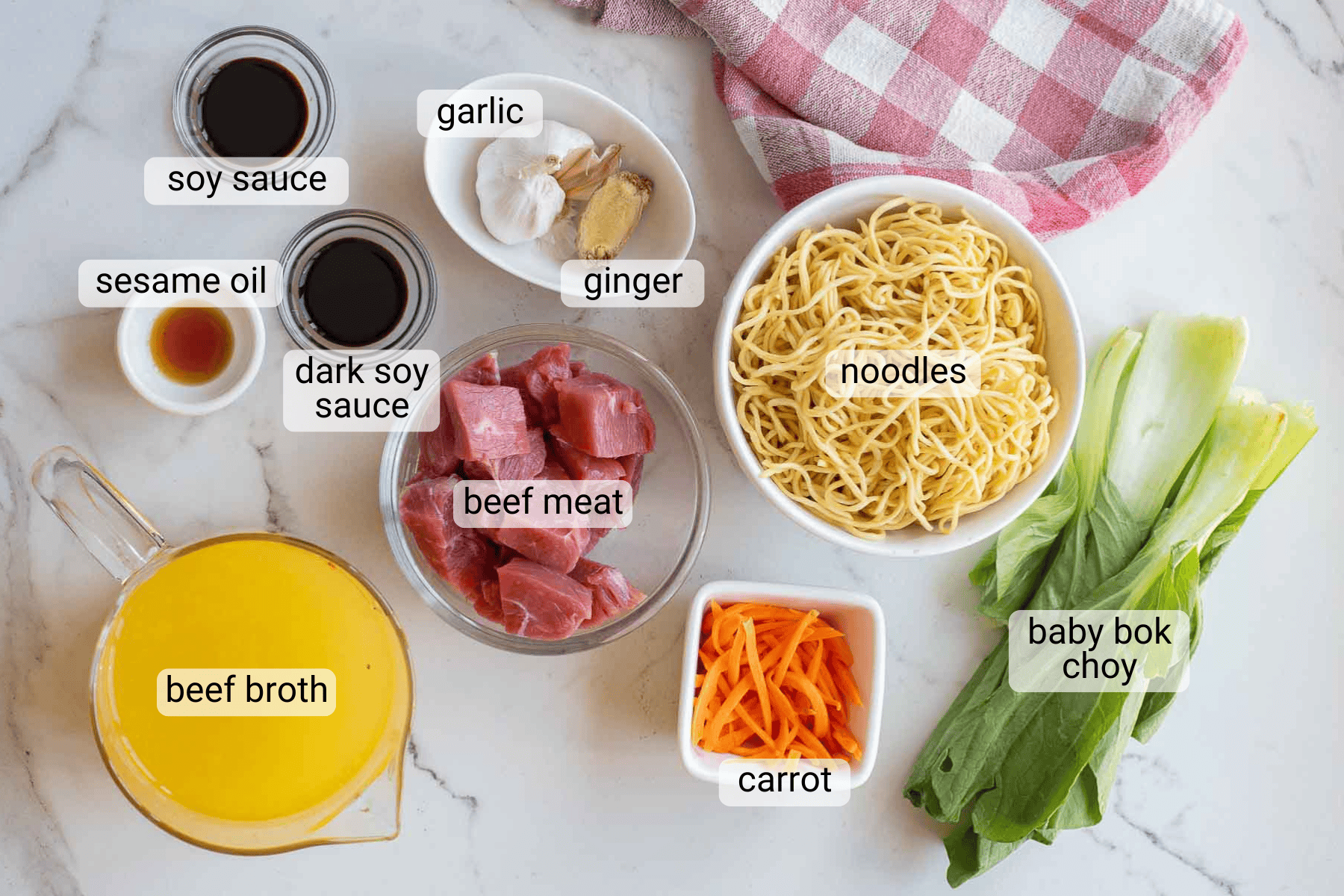 Assorted ingredients for a beef noodle soup are arranged on a counter, including beef meat, noodles, baby bok choy, carrot, beef broth, garlic, ginger, soy sauces, and sesame oil.