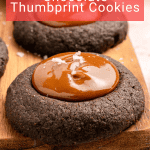 Three chocolate thumbprint cookies with glossy caramel centers and a sprinkle of sea salt, arranged on a wooden board.