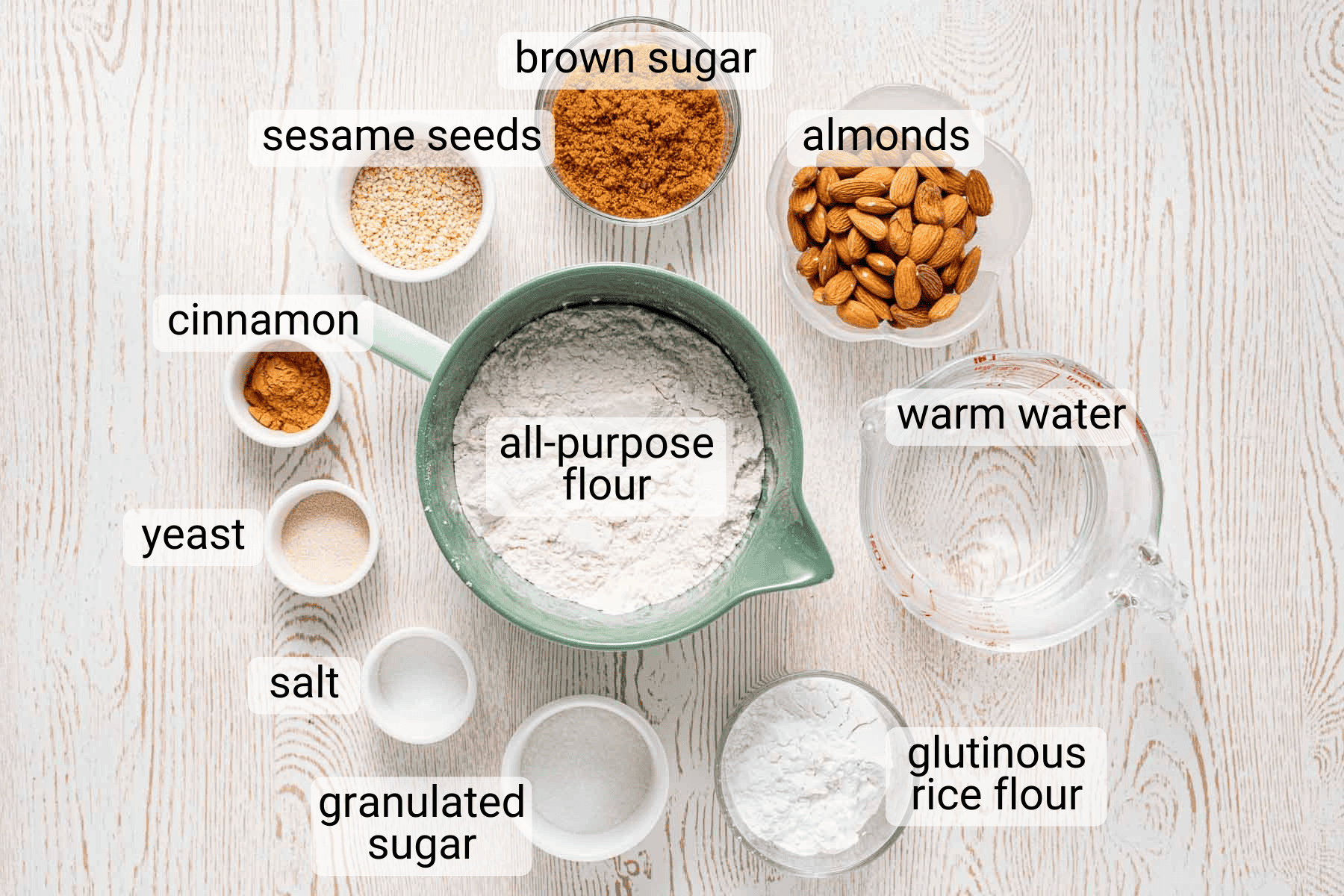 Various labeled ingredients including all-purpose flour, brown sugar, almonds, warm water, glutinous rice flour, granulated sugar, salt, yeast, cinnamon, and sesame seeds on a wooden surface.