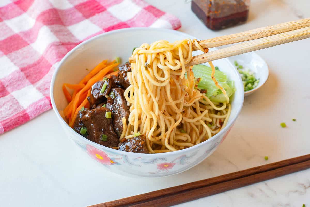 A bowl of noodles with beef, carrots, and greens, with chopsticks lifting noodles, placed on a white surface near a red and white checkered cloth.