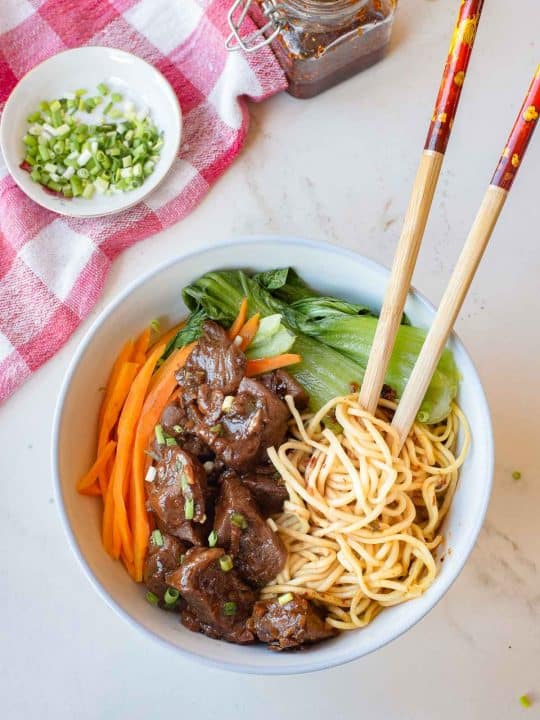 A bowl of noodles with sliced beef, bok choy, and julienned carrots, garnished with chopped green onions, with chopsticks resting on the bowl.