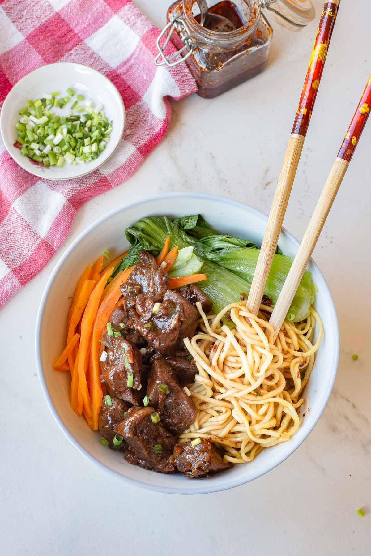 A bowl of noodles with sliced beef, bok choy, and julienned carrots, garnished with chopped green onions, with chopsticks resting on the bowl.