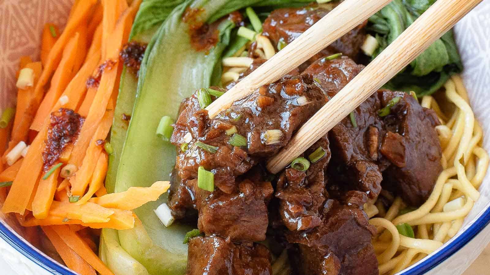 Close-up of chopsticks holding pieces of braised beef over noodles, with bok choy, julienned carrots, and green onions in a bowl.