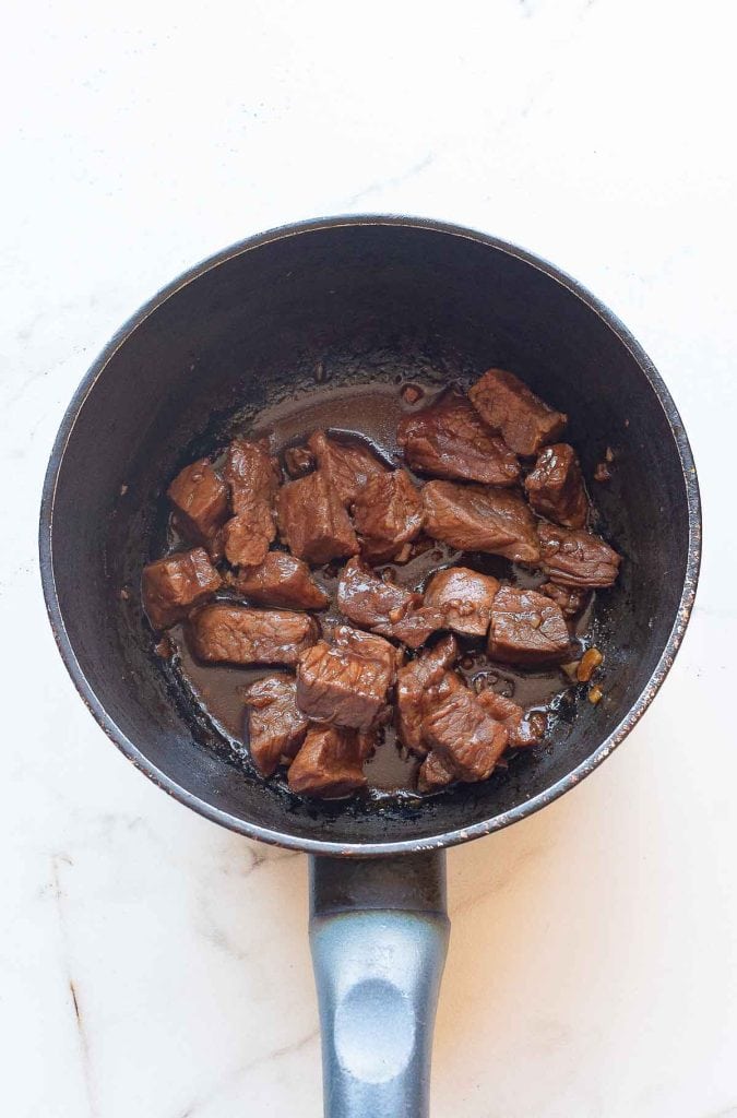 Chunks of cooked beef in a black saucepan on a white marble surface.