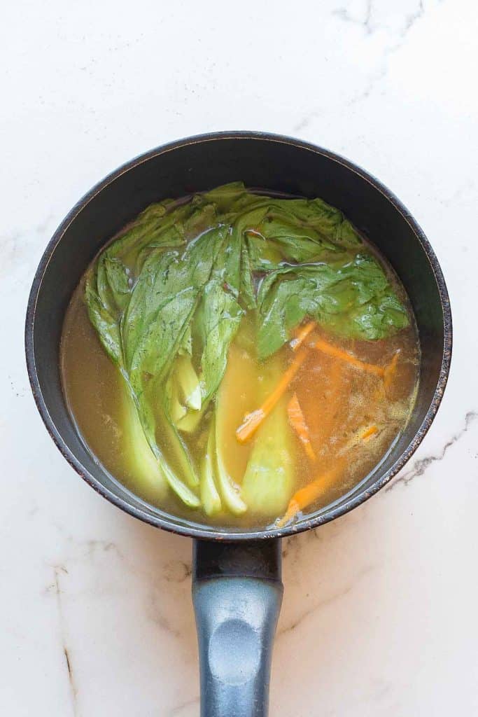 A saucepan filled with broth, bok choy leaves, and sliced carrots on a white marble surface.