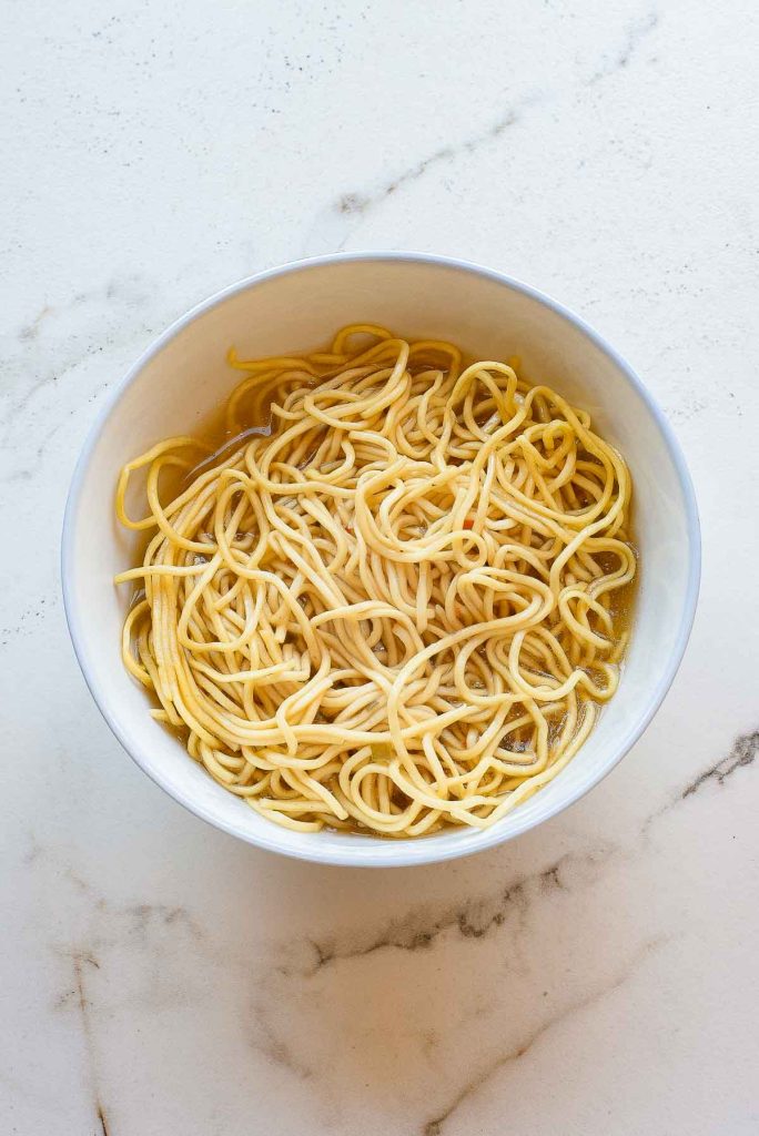 A white bowl filled with plain cooked noodles, placed on a white marble surface.