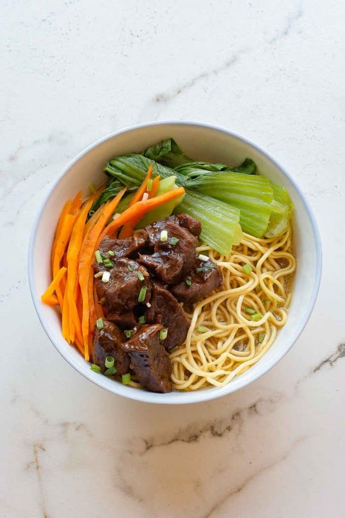 A bowl of noodle soup topped with braised beef, sliced carrots, bok choy, and chopped green onions on a light-colored surface.