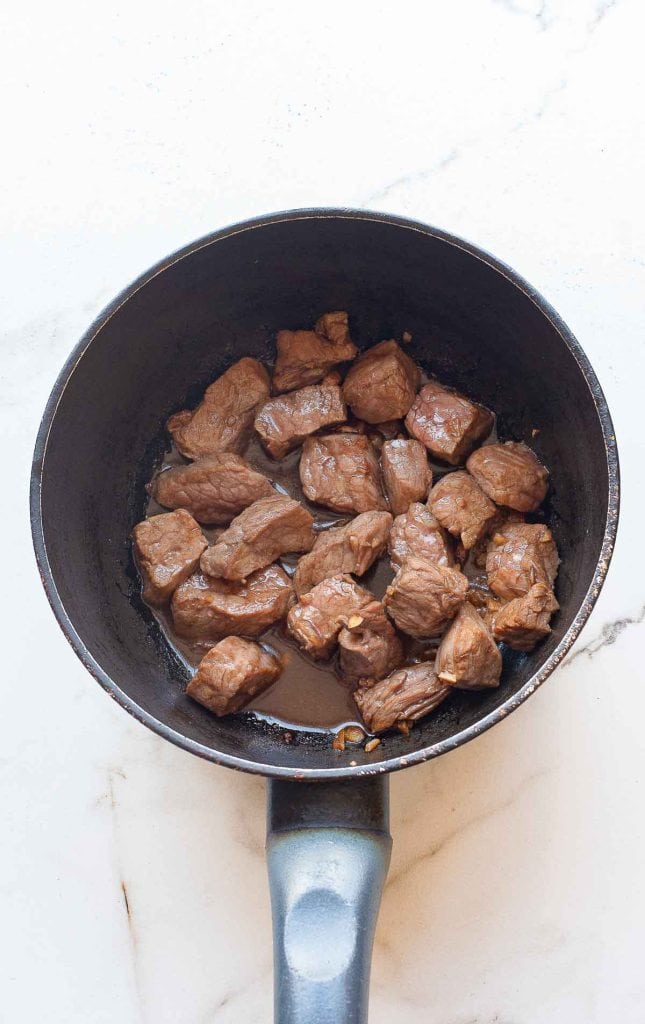 A saucepan containing cooked chunks of beef on a white marble surface.