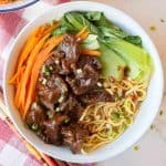 A bowl of noodles topped with sliced beef, bok choy, julienned carrots, and chopped green onions, placed on a red and white checkered cloth.