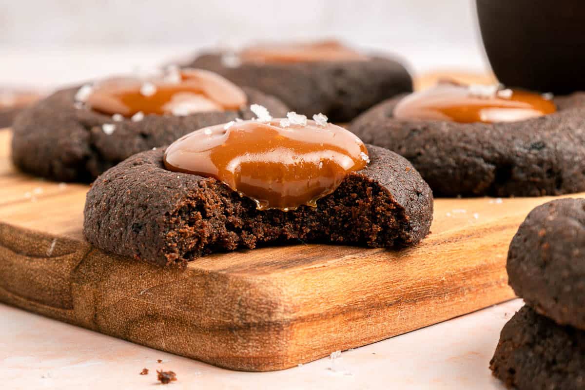 A close-up of a chocolate cookie with caramel topping and a bite taken out, sitting on a wooden board with more cookies in the background.
