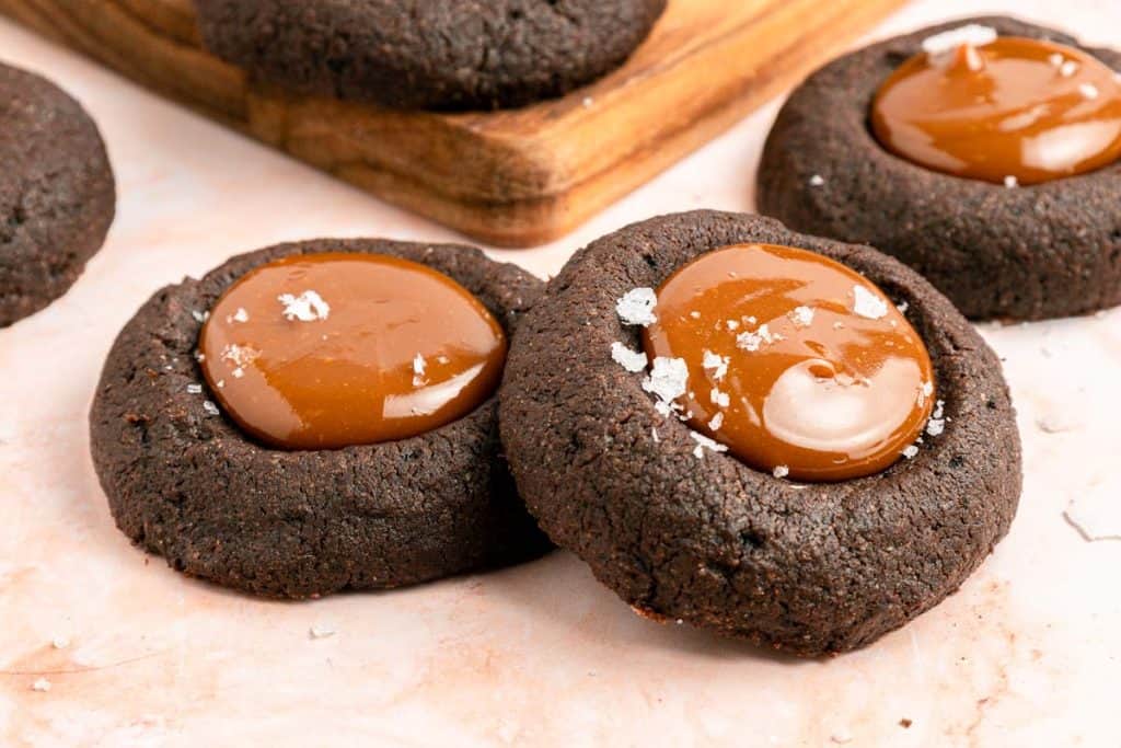 Two chocolate thumbprint cookies filled with caramel and sprinkled with sea salt are placed on a light surface, with more cookies visible in the background.
