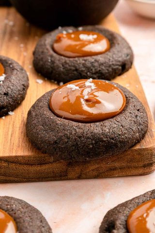 Chocolate thumbprint cookies filled with caramel and topped with a sprinkle of sea salt, arranged on a wooden board.