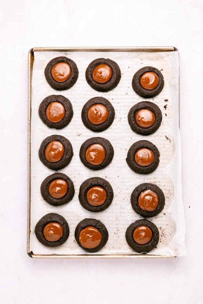 A baking sheet with 15 chocolate thumbprint cookies, each topped with a round dollop of melted chocolate, on parchment paper.