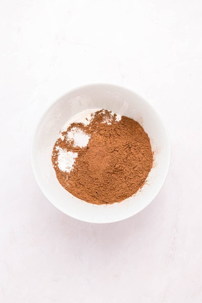 A white bowl containing a mixture of cocoa powder and a small amount of white powder on a light surface.